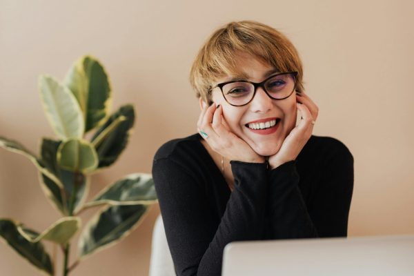 Cheerful woman in eyeglasses and black sweater smiling while working at a desk with a laptop indoors.