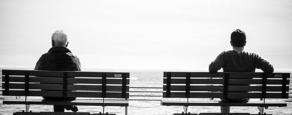 Two people sit on separate benches by the sea, captured in black and white.